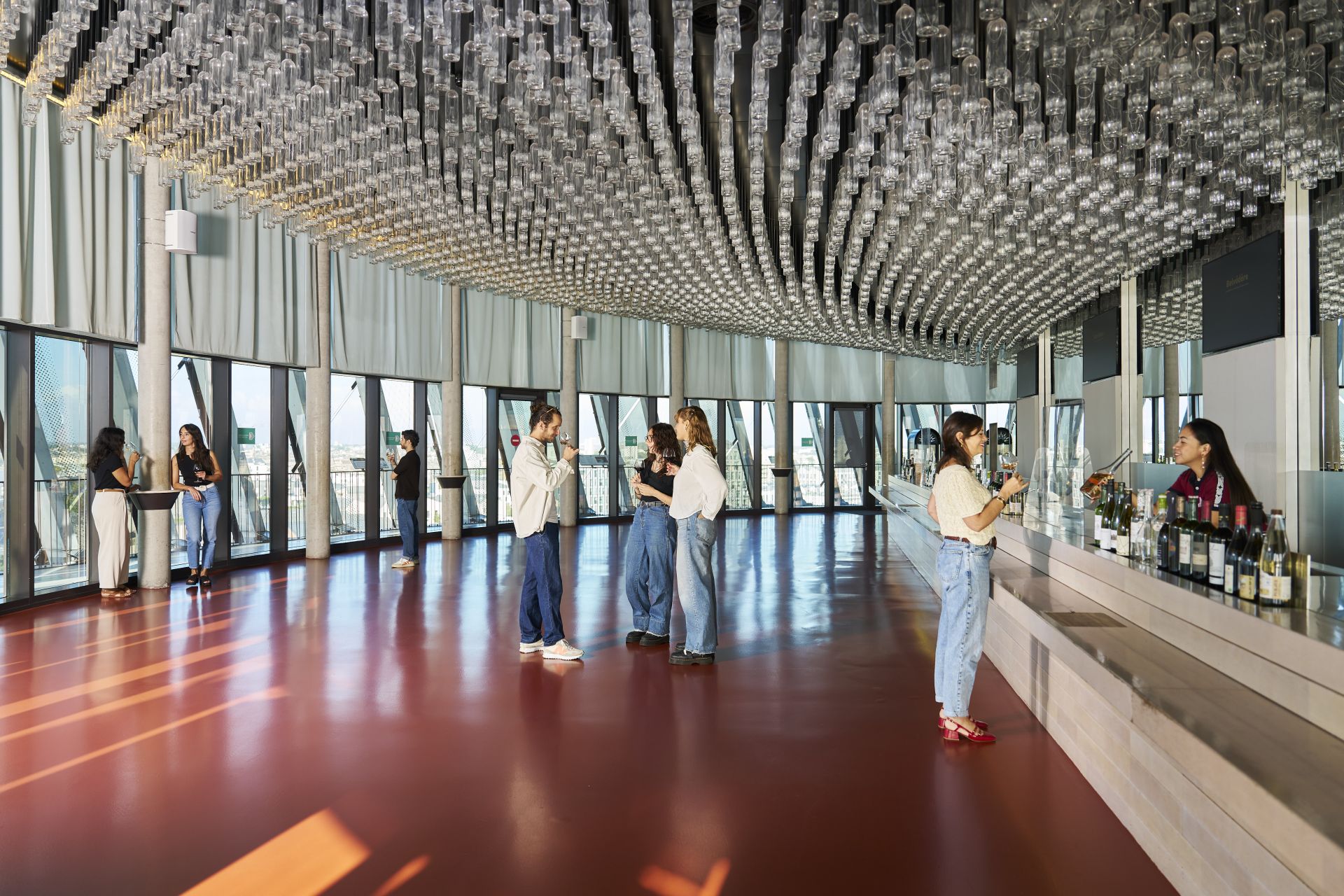 Modern interior space at La Cité du Vin in Bordeaux with visitors exploring the wine museum. The room features a striking ceiling with geometric crystalline glass installations, floor-to-ceiling windows with city views, a deep burgundy floor, and a servic
