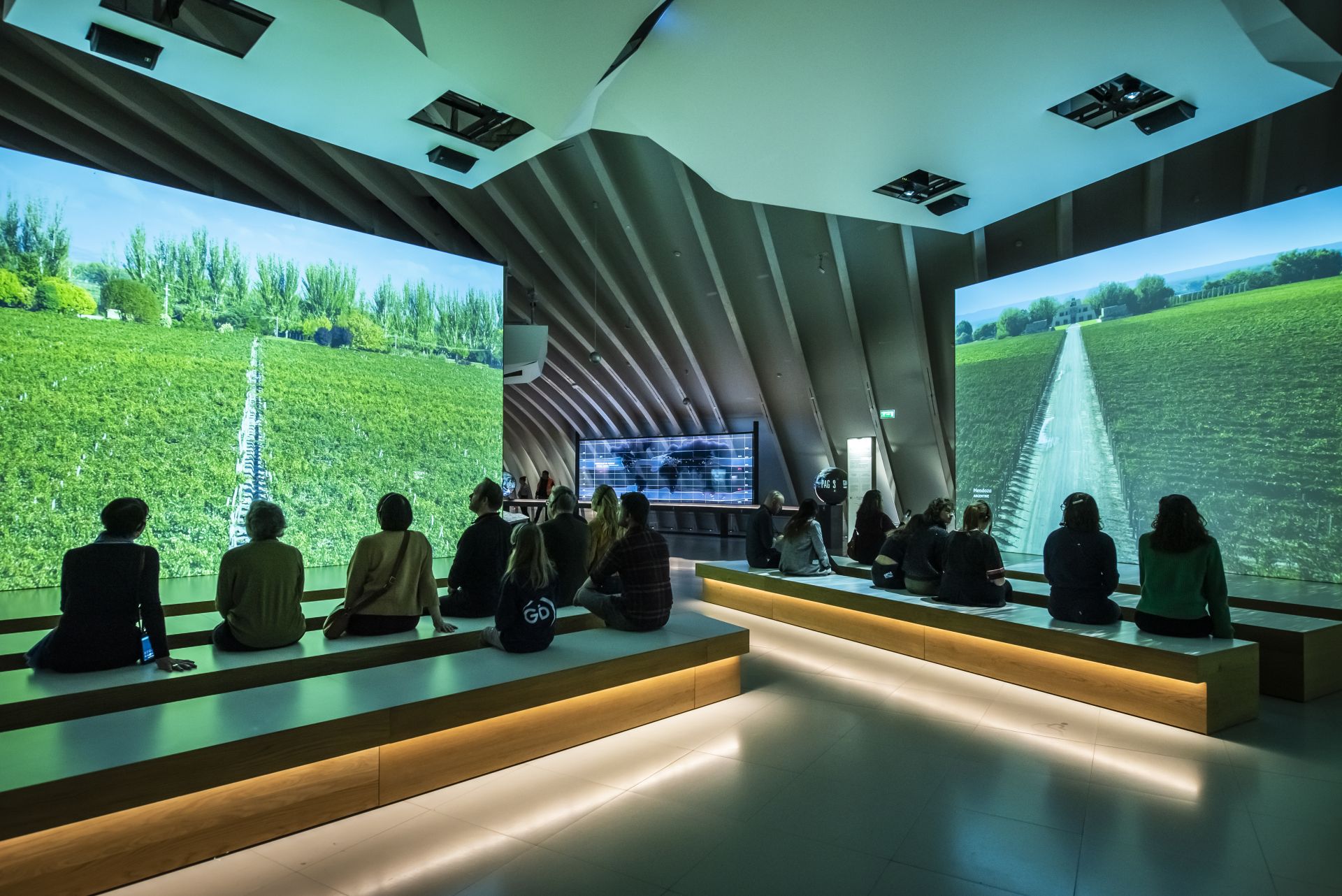 Interior of a modern exhibition space with visitors seated on illuminated benches facing large screens displaying vineyard landscapes, with dramatic angular ceiling architecture and ambient lighting.