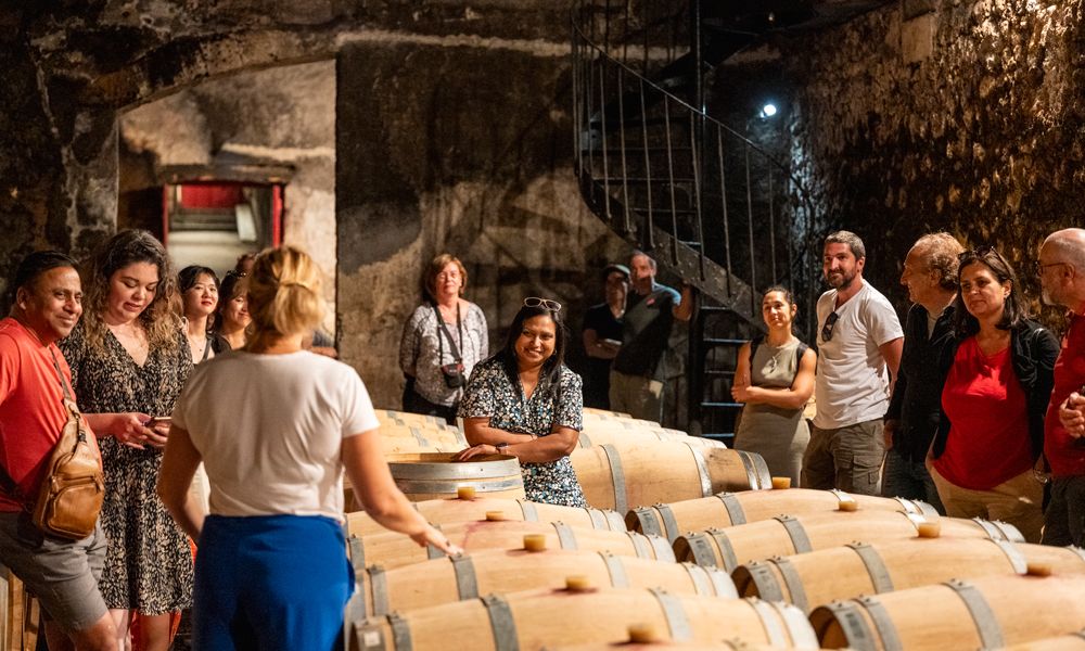 A group of tourists on a guided wine cellar tour in Bordeaux, standing around wooden barrels in an underground stone cellar, with a female guide explaining the winemaking process.