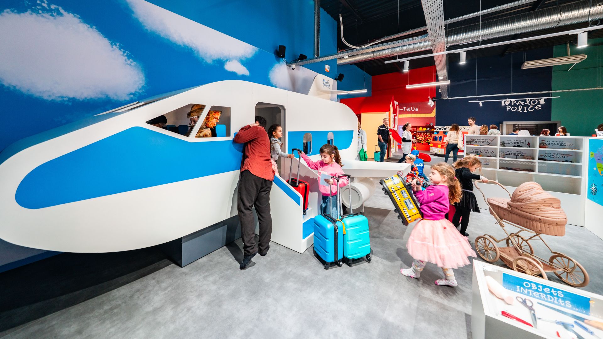 Interactive airplane exhibit at a children's museum in Bordeaux, with kids boarding a white and blue aircraft model, vintage bicycles and suitcases displayed nearby, and various activity stations in the background.