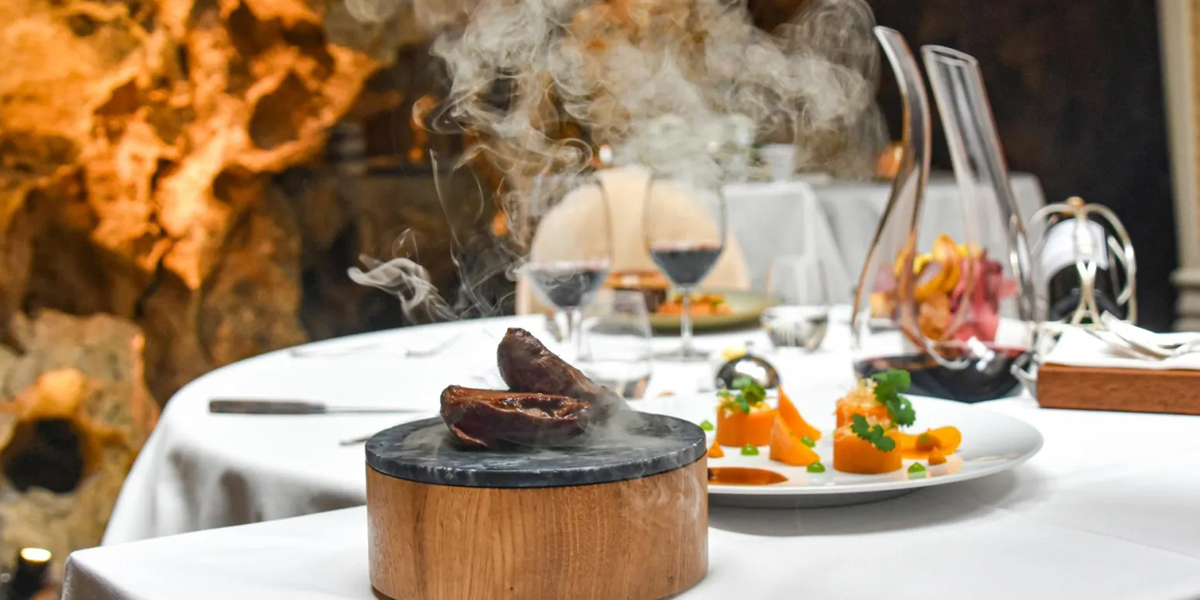 Fine dining presentation with a wooden and slate vessel containing seared meat with smoke, accompanied by elegantly plated vegetables and garnishes on white plates, wine glasses, and refined table setting in an upscale restaurant