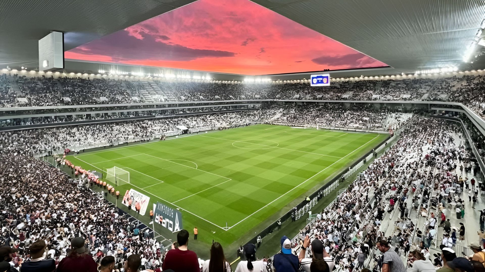 Aerial view of Stade Atlantique in Bordeaux filled with spectators during a rugby match, with a dramatic sunset sky displayed on the large overhead screen and the green pitch ready for play.