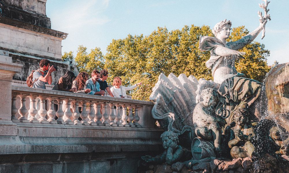 A ornate stone fountain with classical sculptures and water features, featuring a winged figure on a pedestal and cherub statues, with tourists observing from a colonnade balcony overlooking autumn foliage in the background.