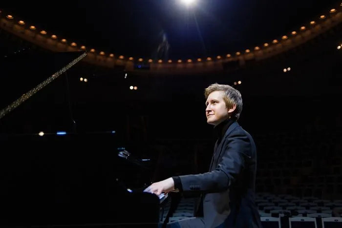 A conductor in a black suit conducting an orchestra in a lit concert hall during a performance of Rachmaninov's Piano Concerto in Bordeaux.