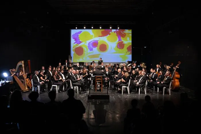 Bordeaux Harmonic Orchestra performing on stage with musicians in formal attire playing various instruments, a projection screen displaying colorful shapes in the background, and a conductor leading the ensemble.