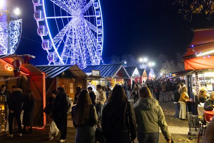 Bordeaux Christmas Market at night featuring an illuminated blue Ferris wheel, decorated wooden chalets, and crowds of visitors browsing market stalls