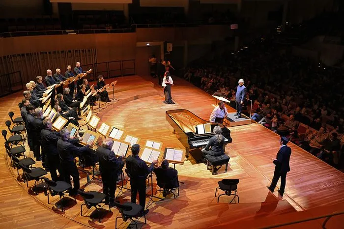 A performance of The Barber of Seville in Bordeaux featuring an orchestra arranged in a semi-circle, solo singers in the center, and an audience seated around the stage in a concert hall.