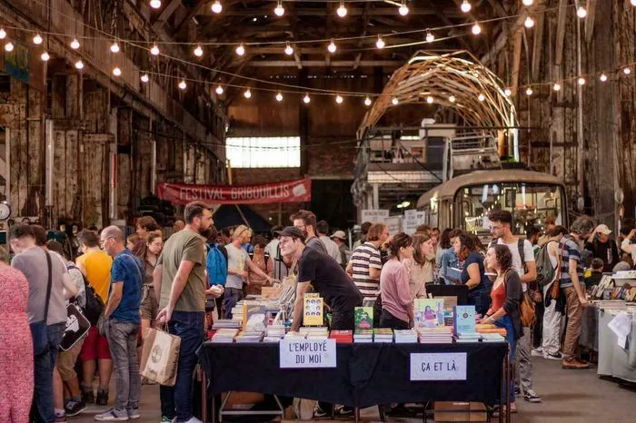Crowd of visitors at Bordeaux's Gribouillis Festival, browsing book and craft vendor stalls in an industrial warehouse decorated with string lights.