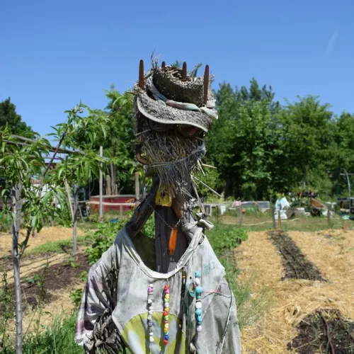 Artistic scarecrow dressed in a white shirt and colorful beads, with a straw head crowned with branches, standing in a community garden in Bordeaux.