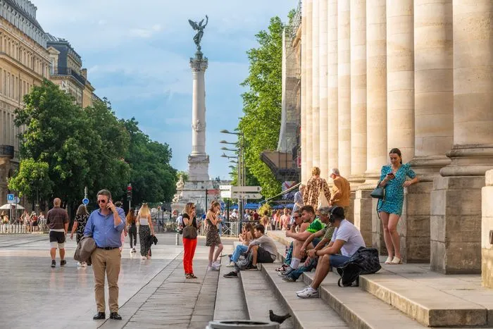 Bordeaux's Place de la Bourse featuring grand neoclassical columns, tourists relaxing on steps, and the Monument aux Girondins monument in the background under blue sky.