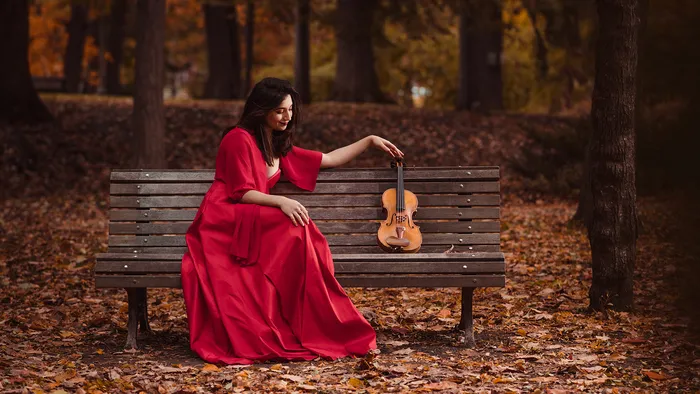 Woman in a red dress sitting on a bench in autumn, holding a violin in a golden leaf forest in Bordeaux