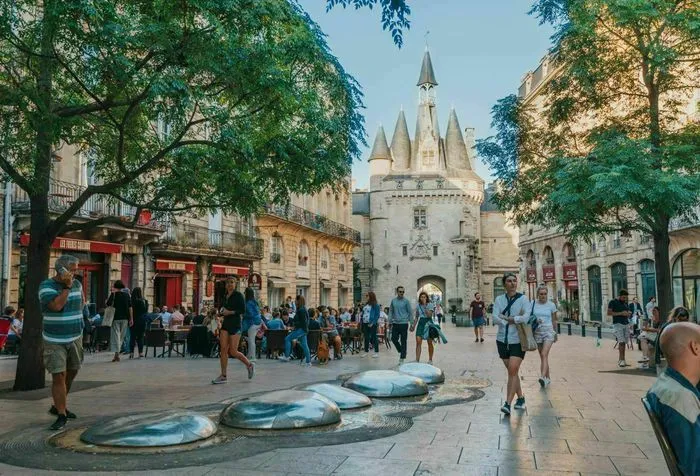 Cailhau Gate, a historic monument in Bordeaux, featuring distinctive pointed towers surrounded by a busy public square with visitors and green trees.