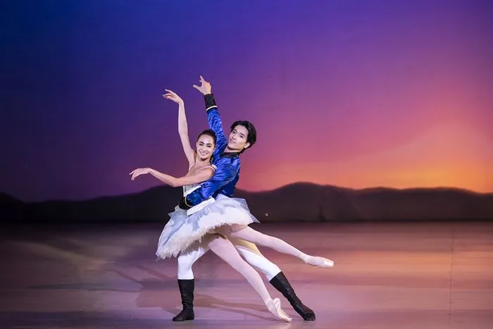 Two ballet dancers in prince and ballerina costumes perform a pas de deux on stage with a sunset backdrop scenery.