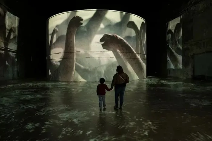 Two visitors, a child and an adult, hold hands in a flooded room at the Bassins des Lumières in Bordeaux, facing giant projections of dinosaurs on the walls and ceiling of the immersive exhibition.