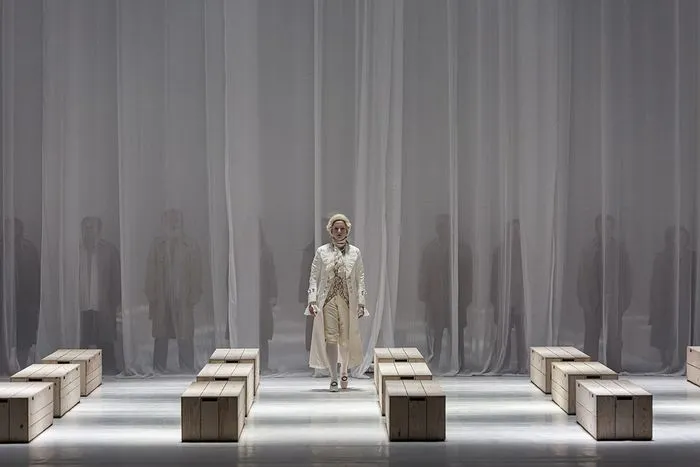 A soloist in light-colored costume and hat stands center stage on a minimalist set with wooden blocks and striped gray curtain backdrop, surrounded by blurred figures, in a production of Mozart's Requiem in Bordeaux.