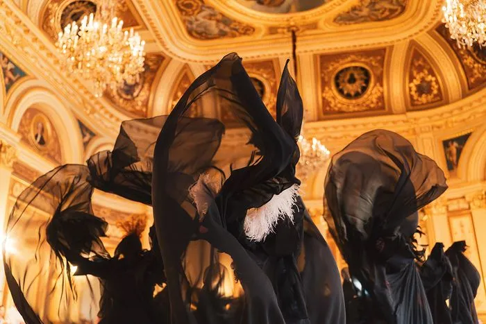 Black-clad figures with veils and pointed hats silhouetted against an ornate gilded ceiling with crystal chandelier at the Witches' Ball in Bordeaux.