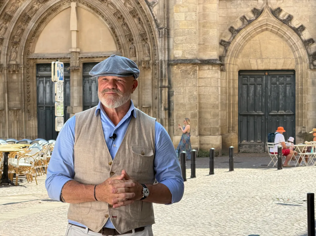 Chef Philippe Etchebest standing in front of a Gothic cathedral on a paved square in Bordeaux, wearing a blue shirt, beige vest, and flat cap.