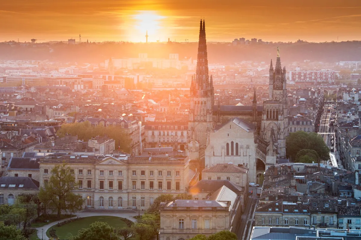 Aerial view of Bordeaux at sunset featuring the Basilica of Saint-Michael and its two Gothic spires in the foreground, overlooking the city's classical architecture and historic districts.