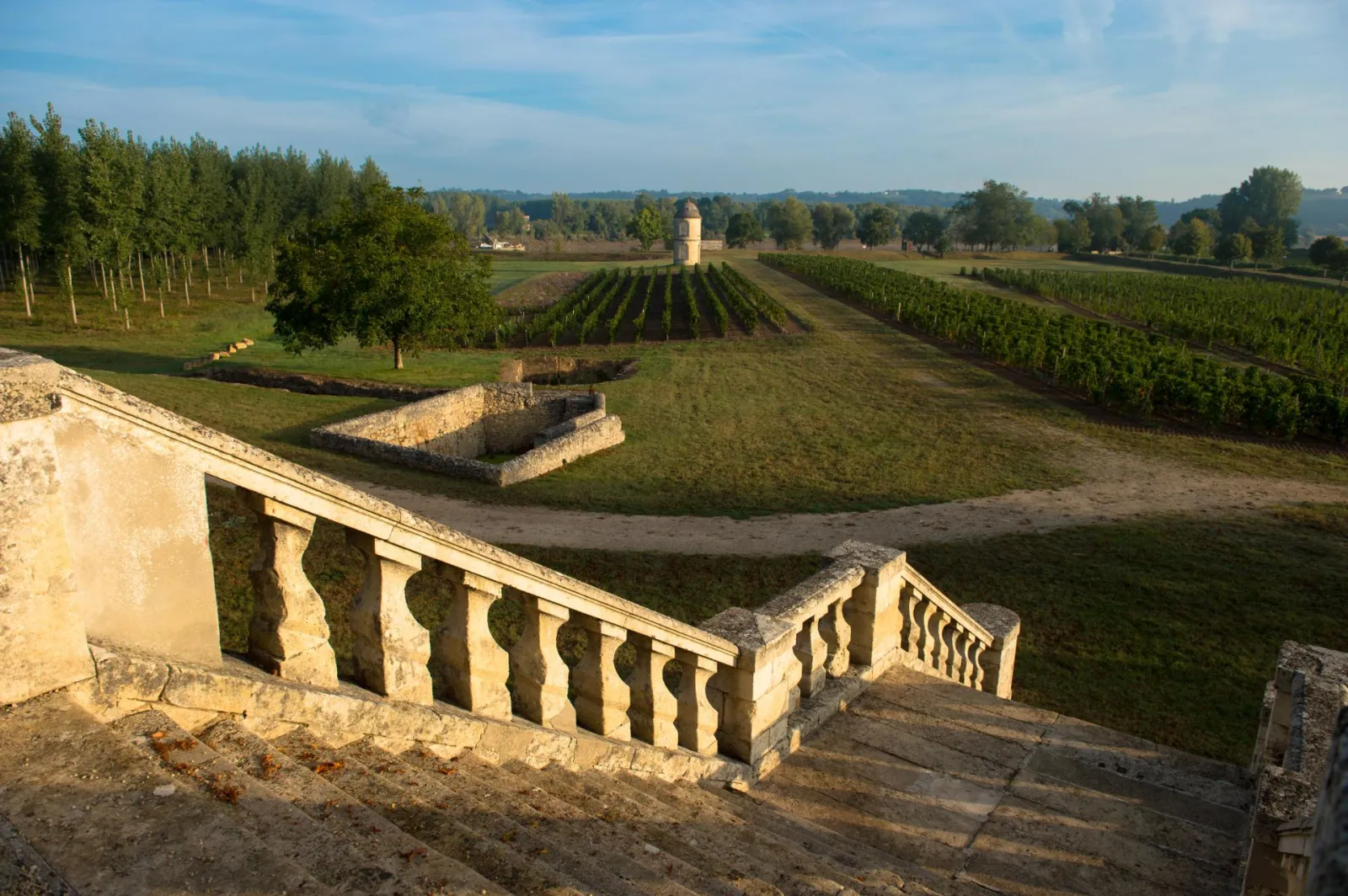 Panoramic view of Château de Portets near Bordeaux featuring stone balustrades, surrounding vineyards, and a stone tower in the background under a blue sky.