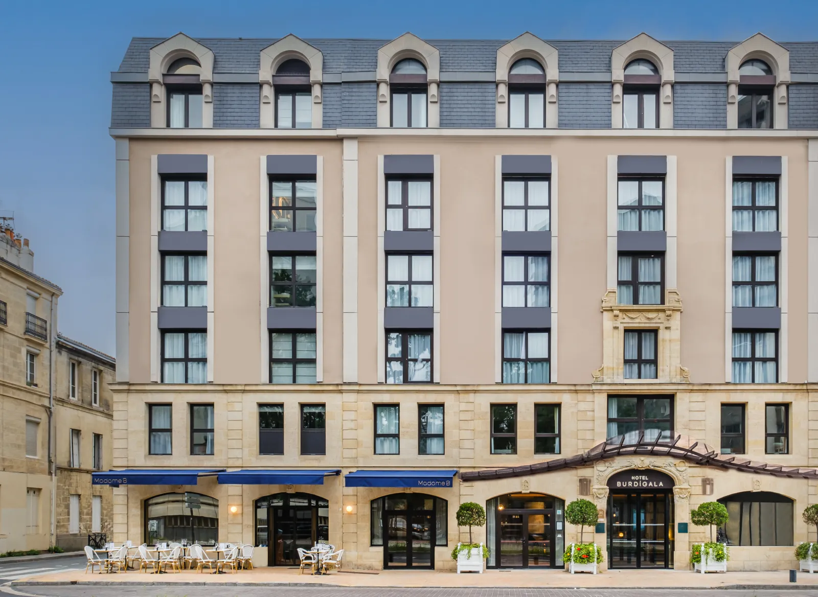 Facade of Hotel Burdigala by Inwood Hotels in Bordeaux, a Haussmann-style building with a commercial ground floor featuring blue storefronts and terrace, topped by six office floors with regular windows and an attic story with arched dormers.