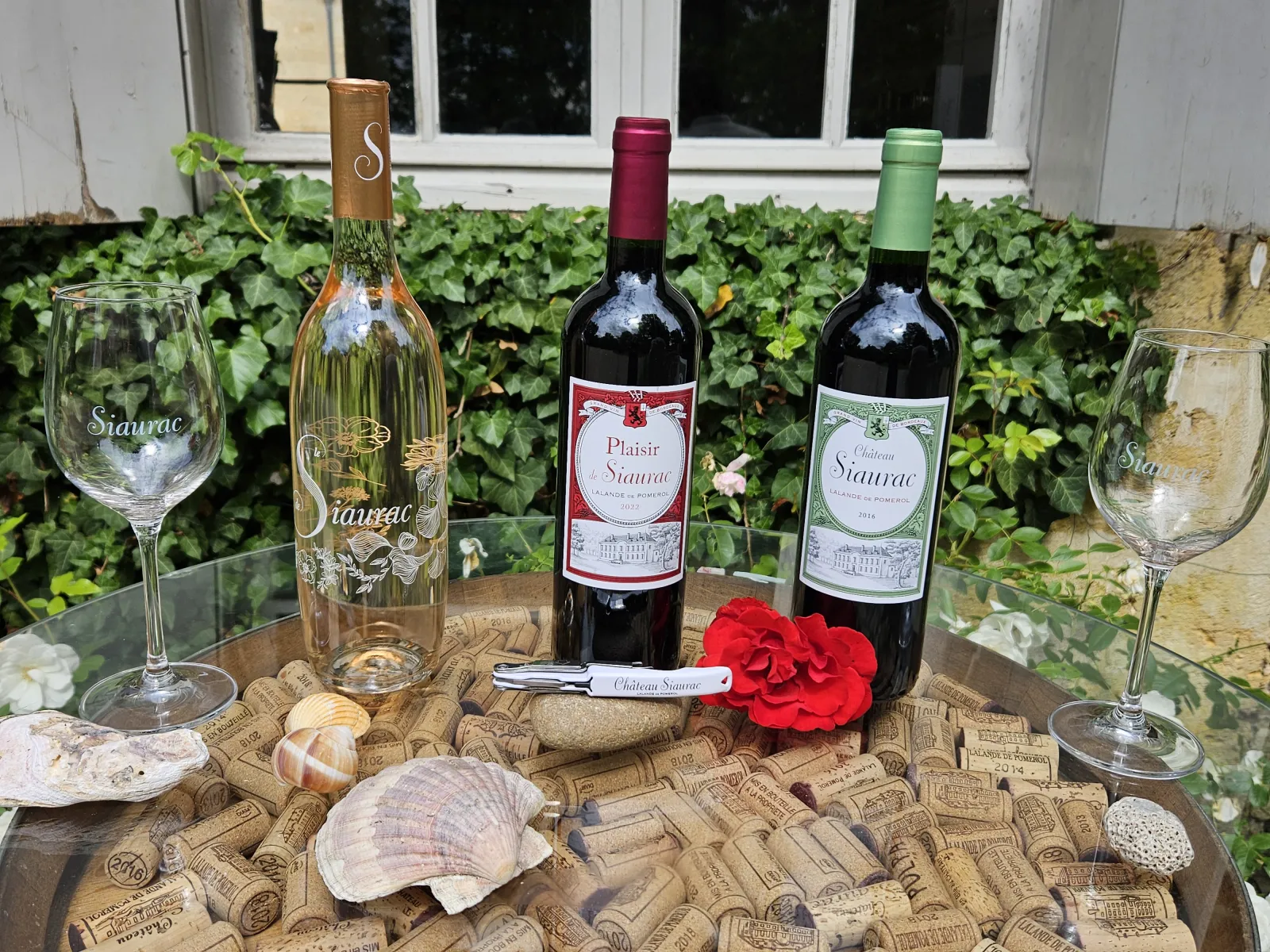 Three bottles of Château Siaurac Bordeaux wine (white, red Plaisir, and red) displayed on a table decorated with wine corks, seashells, and a red flower, accompanied by engraved Siaurac wine glasses, with a flowering garden in the background.