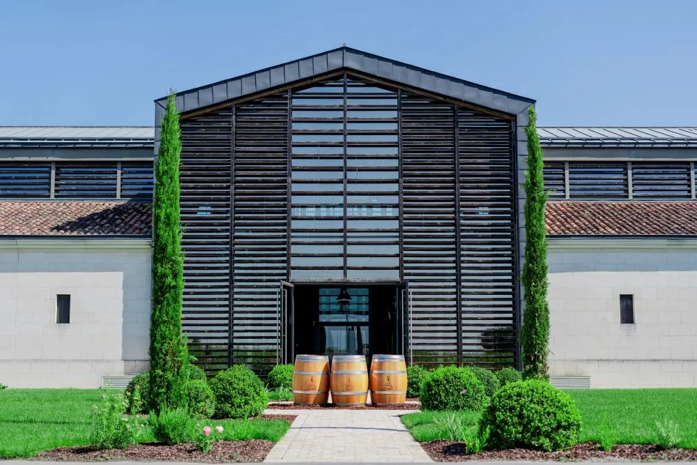 Modern facade of Château La Fleur de Boüard in Bordeaux featuring a black wooden architectural structure, three wooden wine barrels at the entrance, surrounded by greenery and cypress trees.