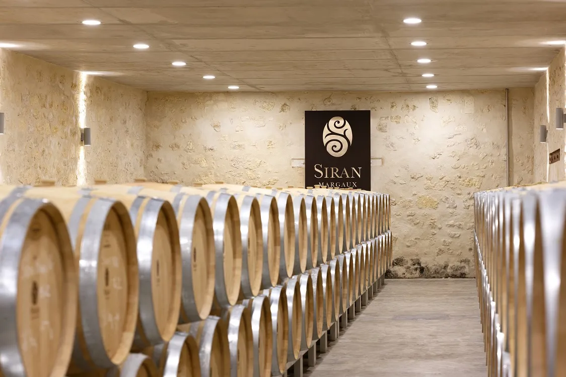 Wine cellar at Château Siran in Bordeaux featuring rows of oak barrels lined symmetrically under recessed lighting, with exposed stone walls and the estate's logo displayed in the background