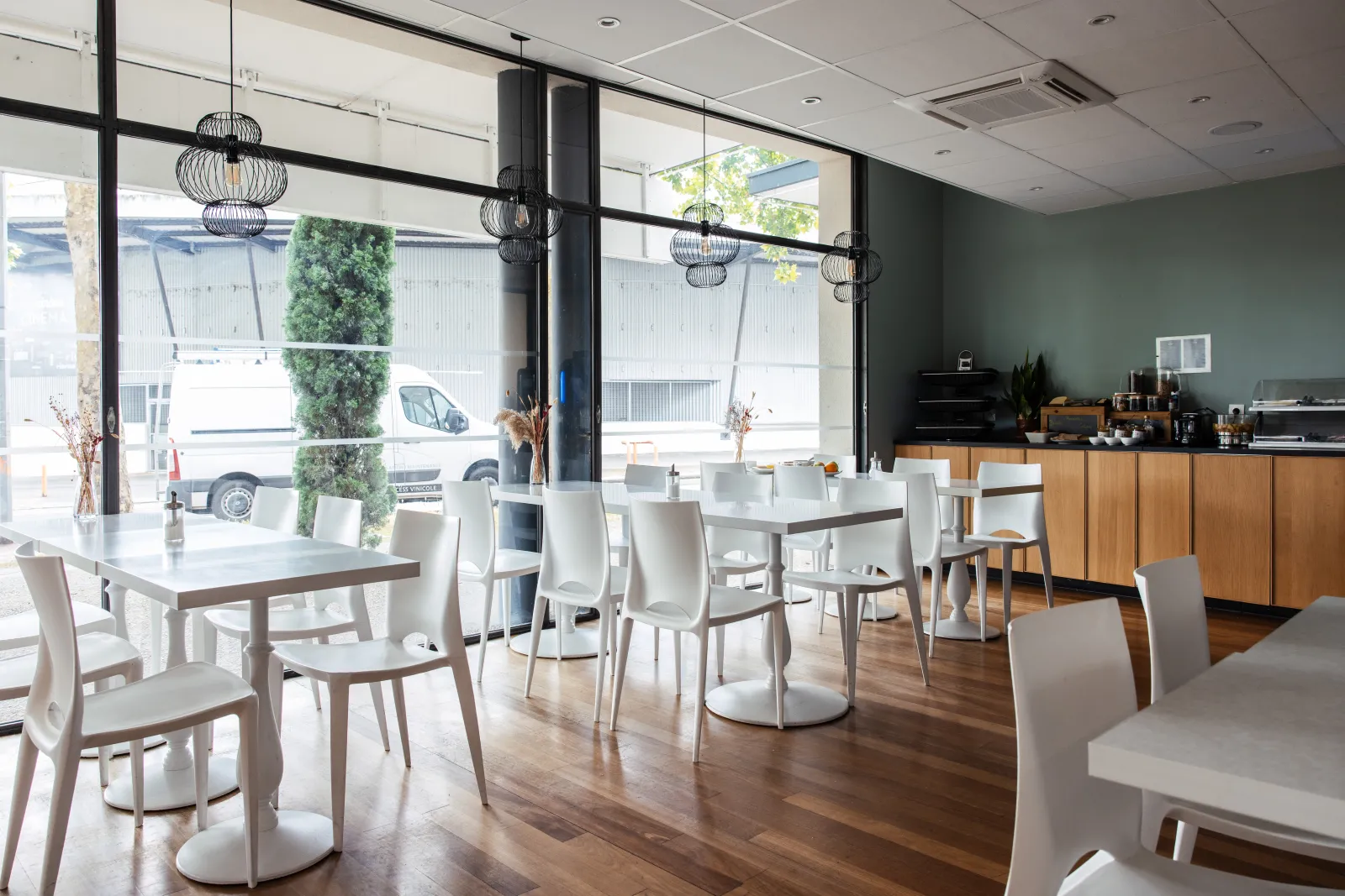 Modern breakfast room with white tables, grey chairs, large windows overlooking the street, black designer pendant lights and equipped kitchen in the background at Talence Centre.