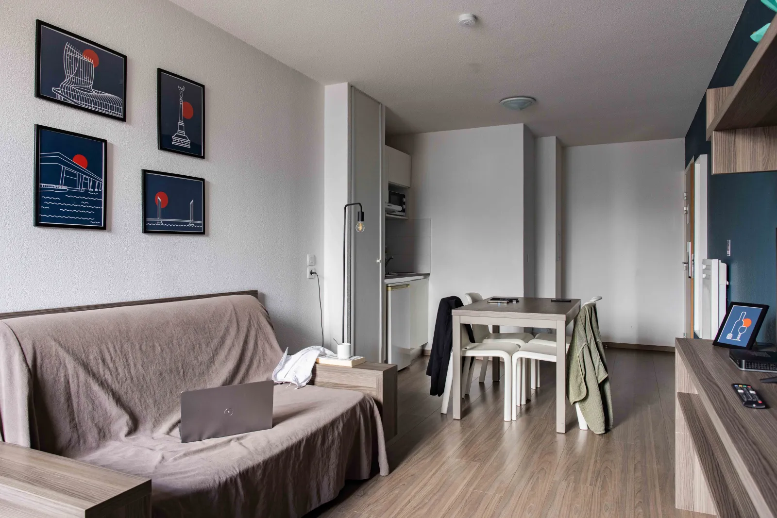 Modern studio at Ténéo Aparthotel Bordeaux-Maritime featuring a gray sofa, white dining table, equipped kitchen, and wall decor with four black frames depicting Bordeaux landmarks.