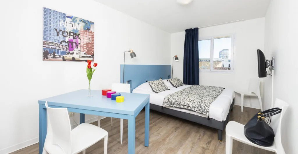Modern bedroom at Ténéo Aparthotel Bordeaux Saint-Jean featuring a double bed, light blue desk, white chair, New York wall art, city-view windows, and wooden flooring.