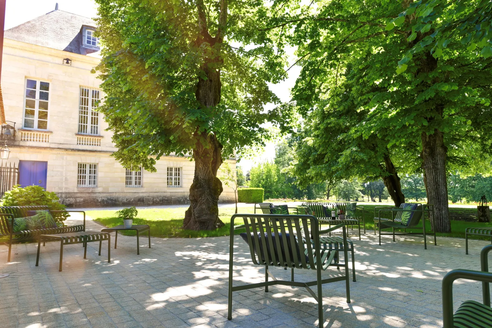 Shaded terrace at Château La Louvière in Bordeaux with green chairs beneath ancient trees, stone château facade visible in the background