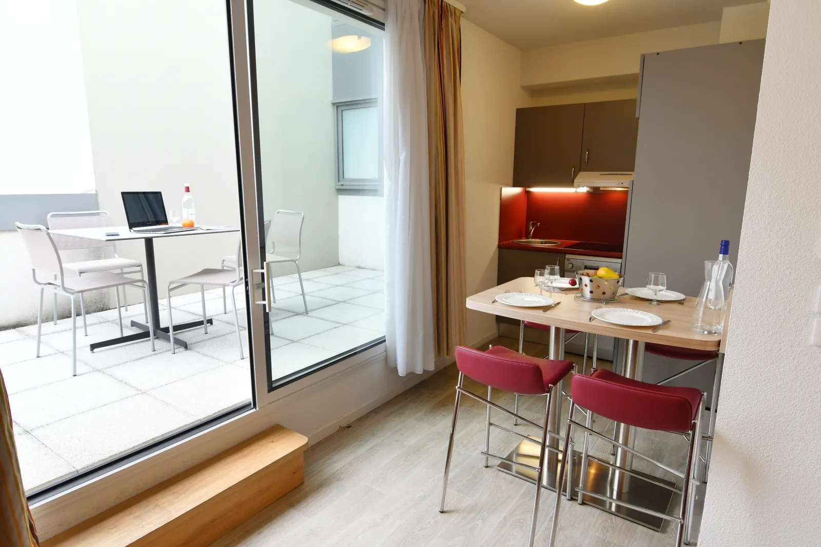 Modern apartment-hotel interior in Bordeaux featuring an equipped kitchen with red backsplash, dining area with two red chairs, wooden table, and access to a balcony with terrace view.
