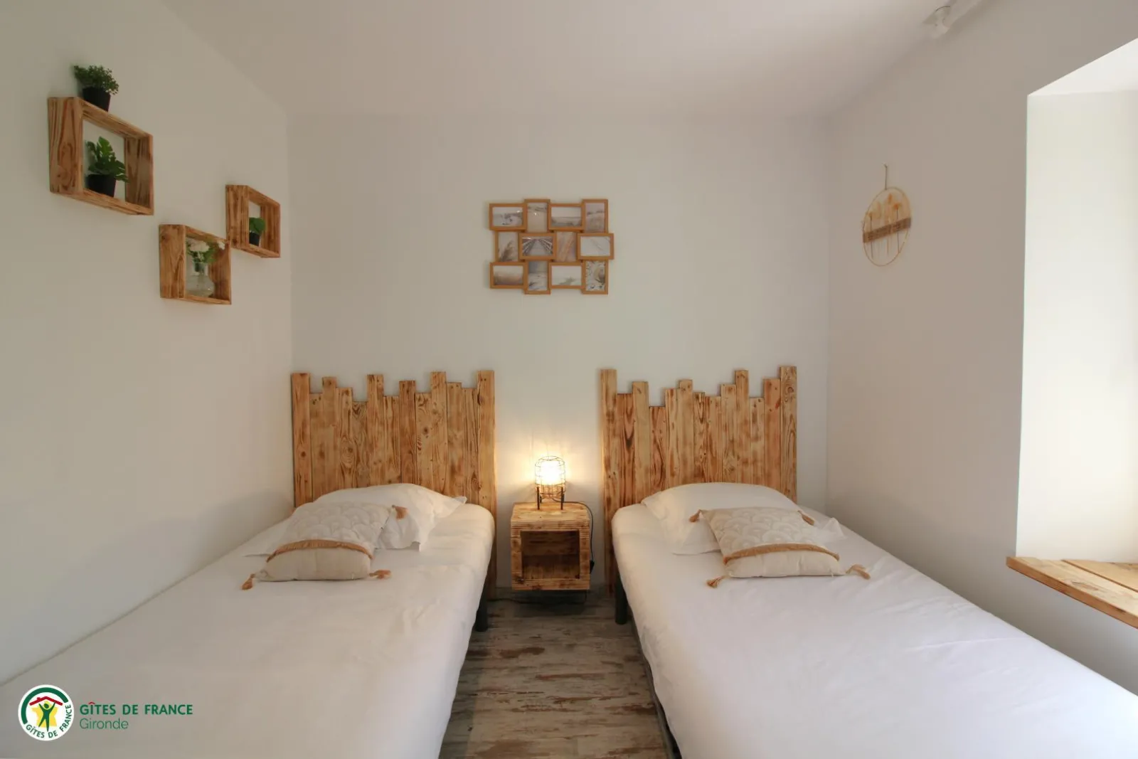 Twin bedroom with two single beds featuring raw wooden headboards, minimalist decor with wooden wall frames and potted plants, wooden nightstand between beds with an illuminated lamp