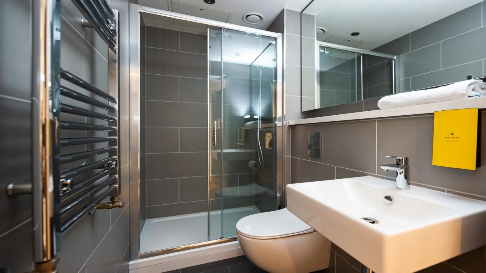 Modern bathroom in Staycity Aparthotel Bordeaux featuring walk-in shower, double sink, toilet, black towel radiator and grey tile walls