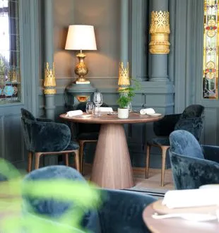 Elegant restaurant dining room featuring a round wooden table, blue velvet chairs, gold floor lamp, gray-blue wall panels with golden accents, at Grand Barrail restaurant in Bordeaux