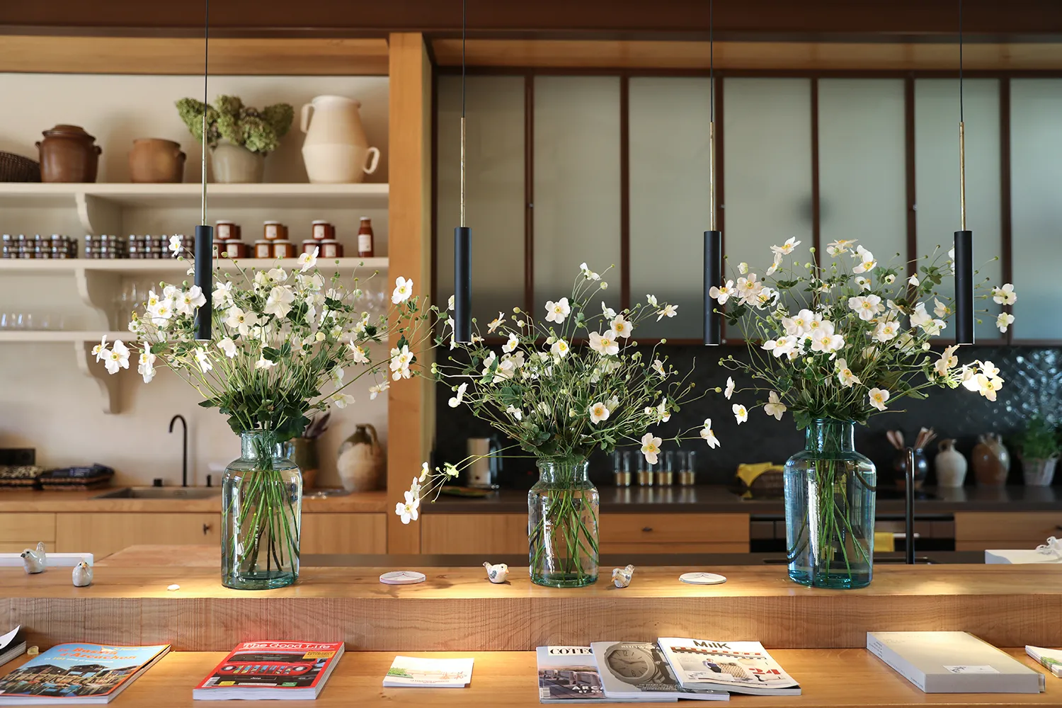 Wooden table featuring three glass vases with white and yellow daisies, black hanging lights above, modern kitchen in the background, magazines on the table, bright and elegant interior.