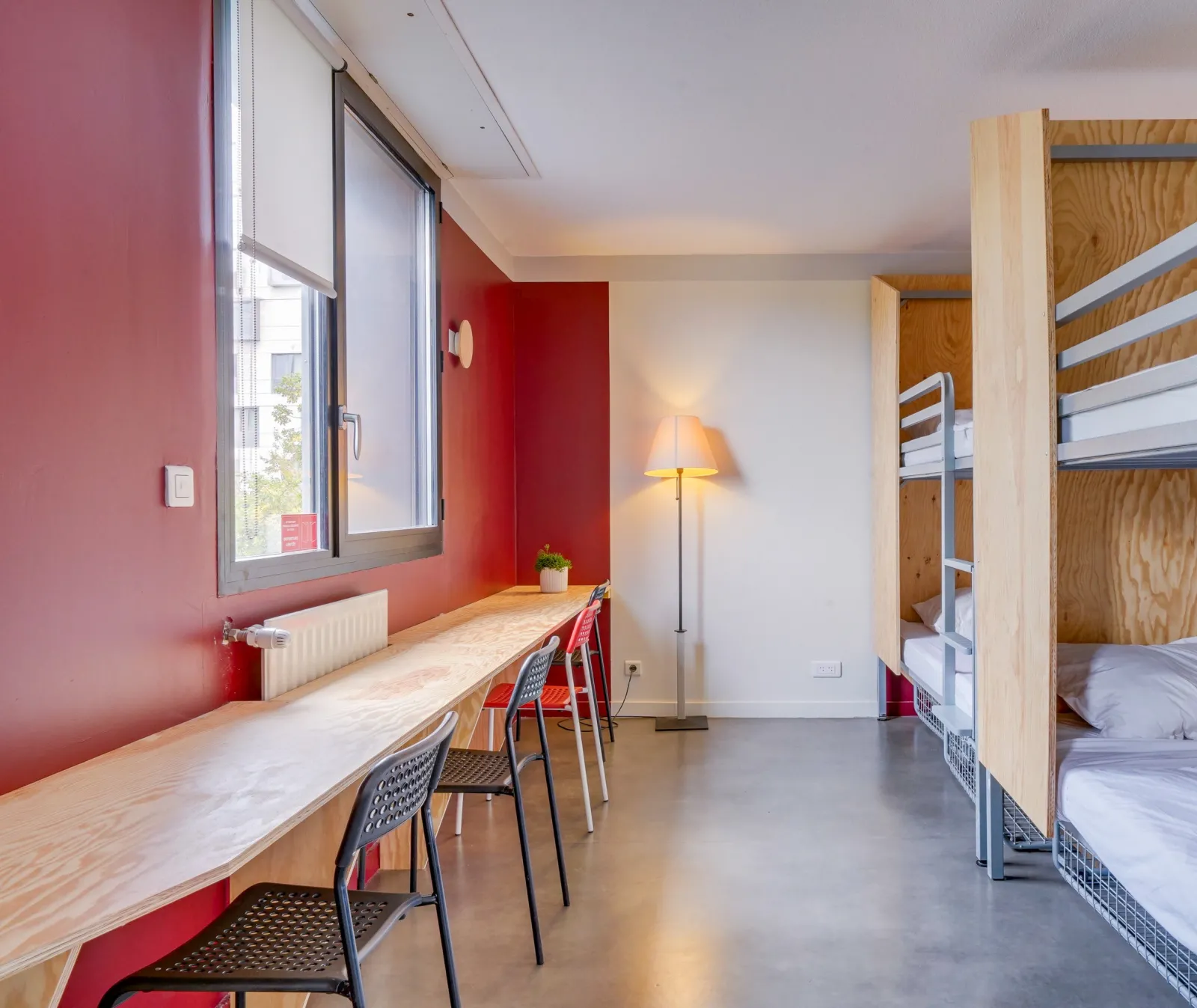 Modern youth hostel room in Bordeaux featuring wooden bunk beds, desk with red chairs, vibrant pink wall, window with view, and standing lamp