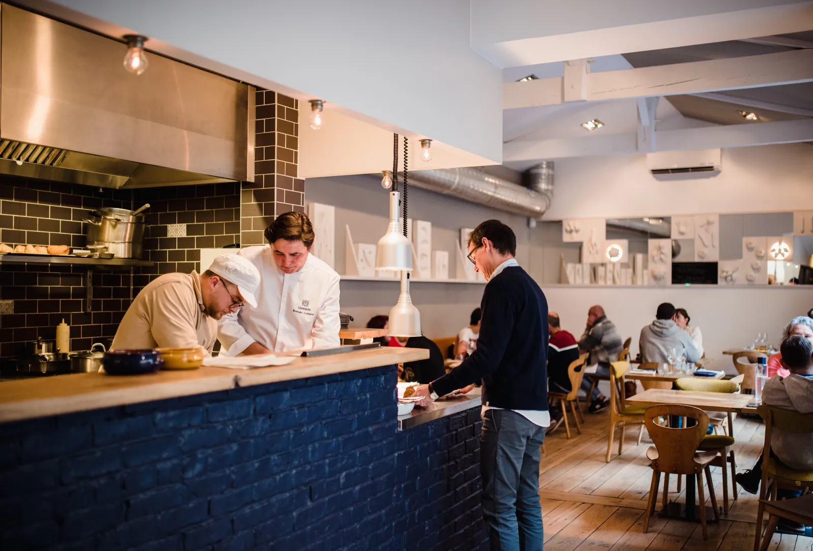 Interior of a Bordeaux restaurant featuring an open kitchen counter where two white-clad chefs serve a customer, modern setting with black brick walls and diners seated in the background.