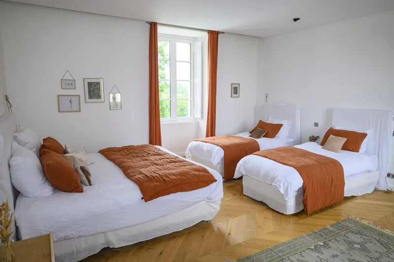 Spacious hotel bedroom with three white beds with orange pillows and throws, orange curtains on window, wooden parquet floor, minimalist wall décor and white radiator