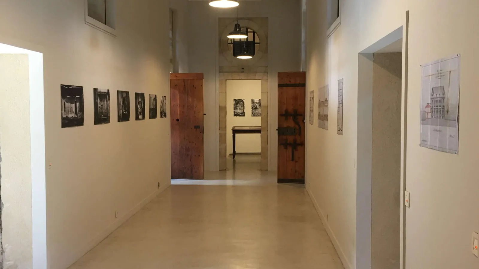 Exhibition corridor at the Bordeaux Museum of Decorative Arts and Design displaying black and white photographs on walls, red wooden doors, and architectural plans.