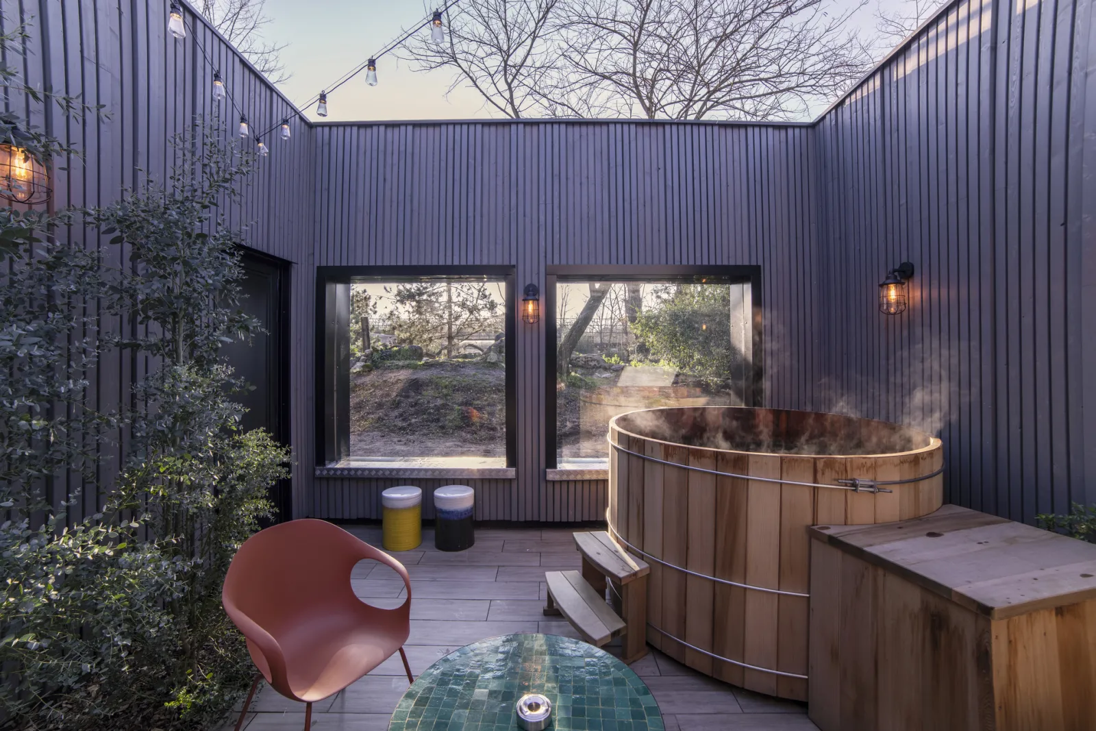 Covered terrace with round wooden hot tub, pink chair and large windows overlooking a wooded garden at dusk in Bordeaux