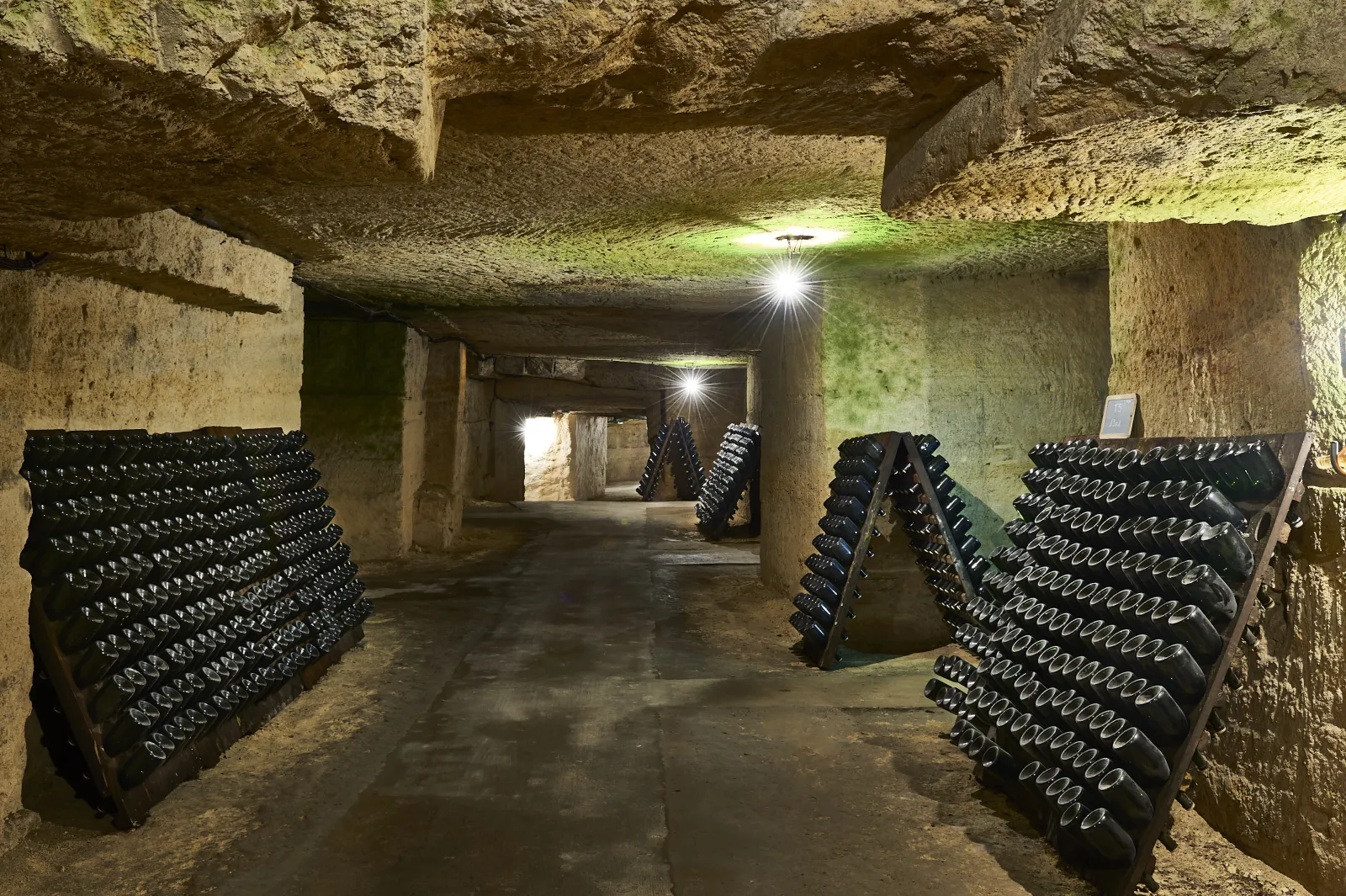 Underground wine cellar in the Cordeliers district of Bordeaux featuring stacked wine bottles on both sides, modern LED lighting, and stone ceiling and walls.