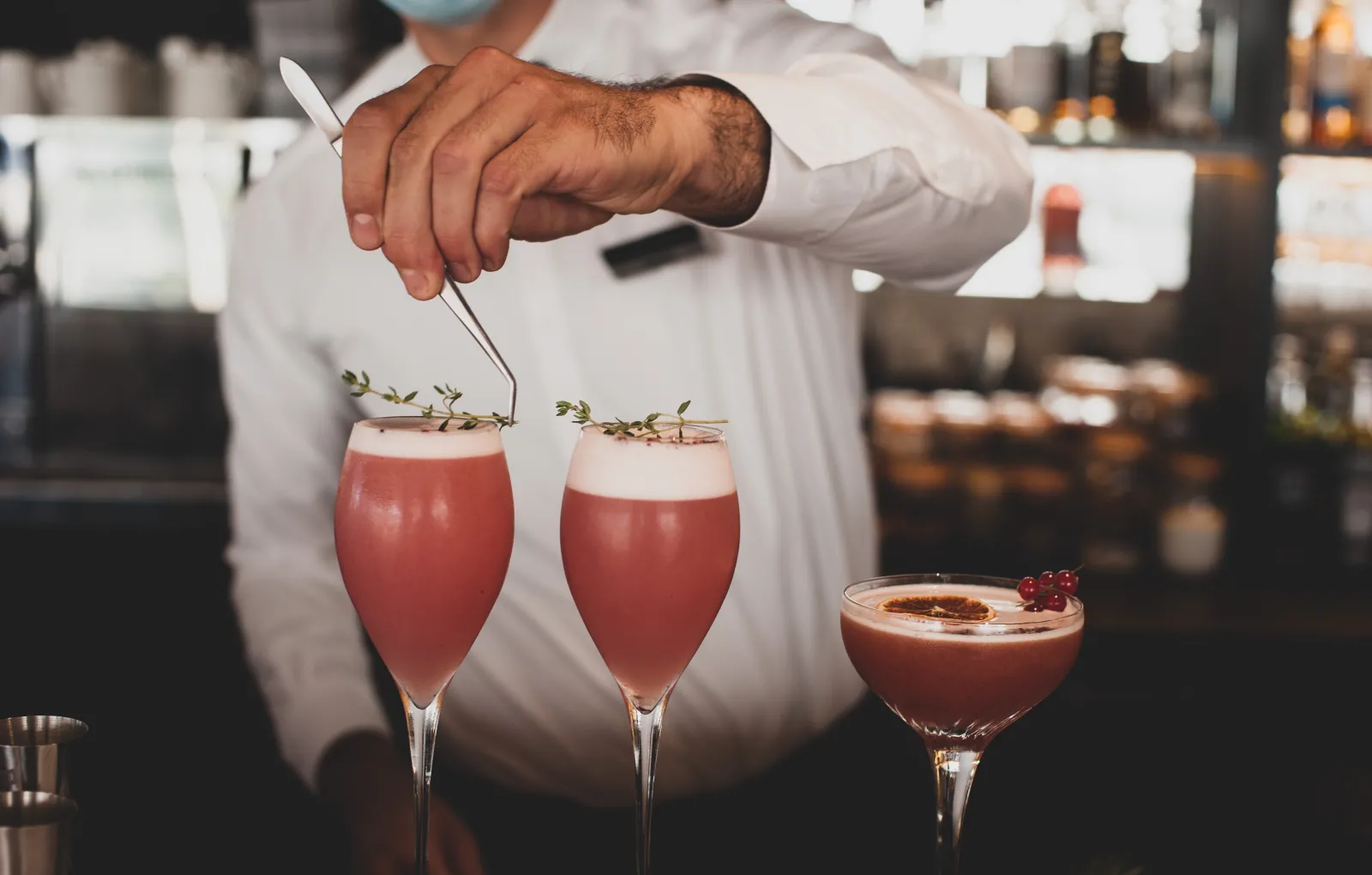 A bartender in white attire crafts two red cocktails garnished with fresh thyme at the Radisson Blu Hotel Bordeaux, with a third glass on the counter