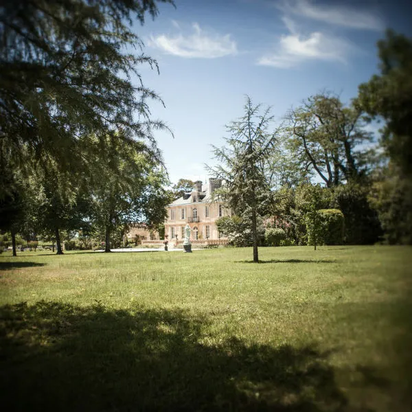 View of Château Kirwan in Bordeaux from its park, featuring a pink stone mansion surrounded by trees and green lawn under a clear sky.