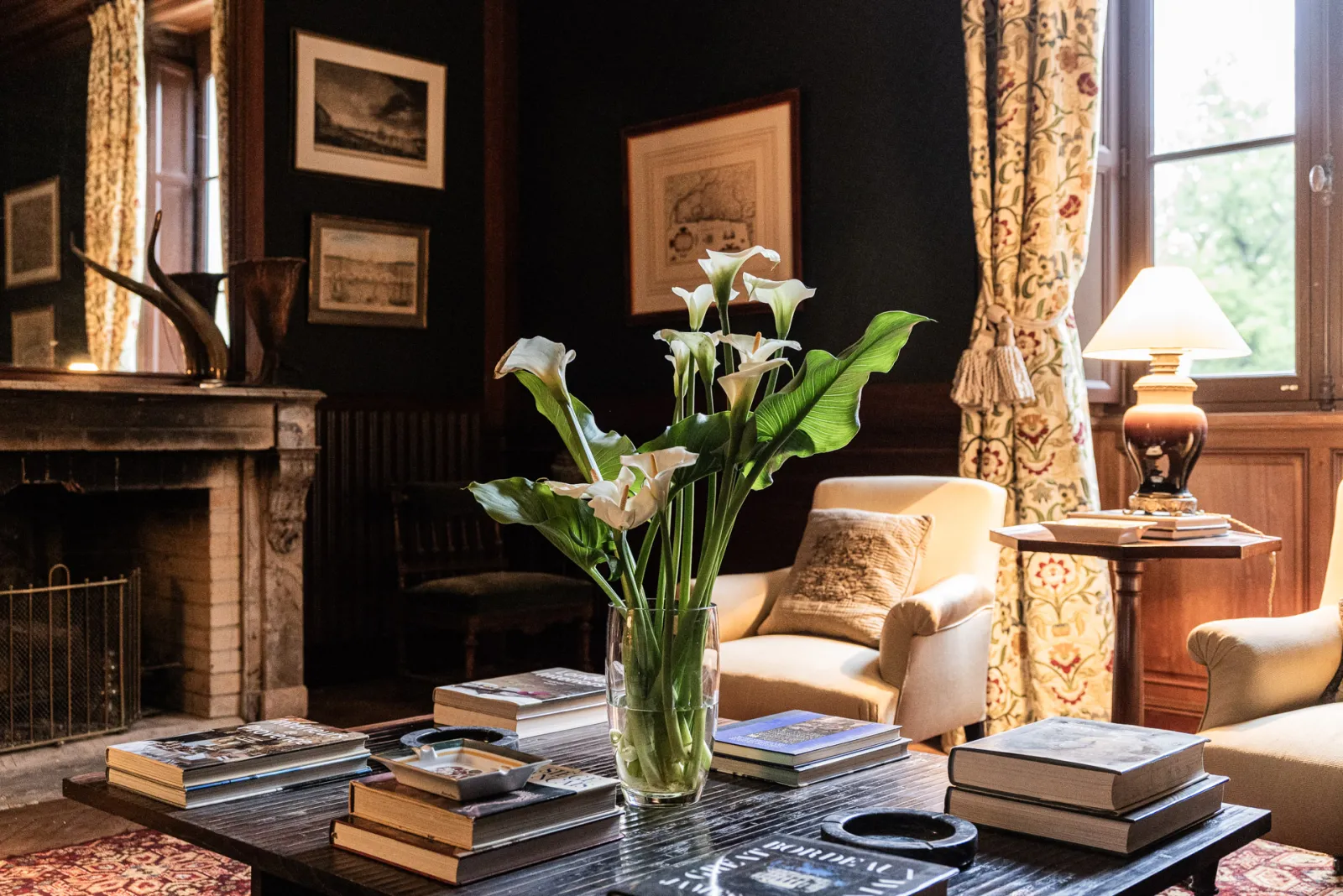 Elegant living room at Château Sénéjac in Bordeaux featuring white calla lilies in a vase on a coffee table surrounded by books, cream armchairs, fireplace, gold curtains, and black walls with framed artwork.