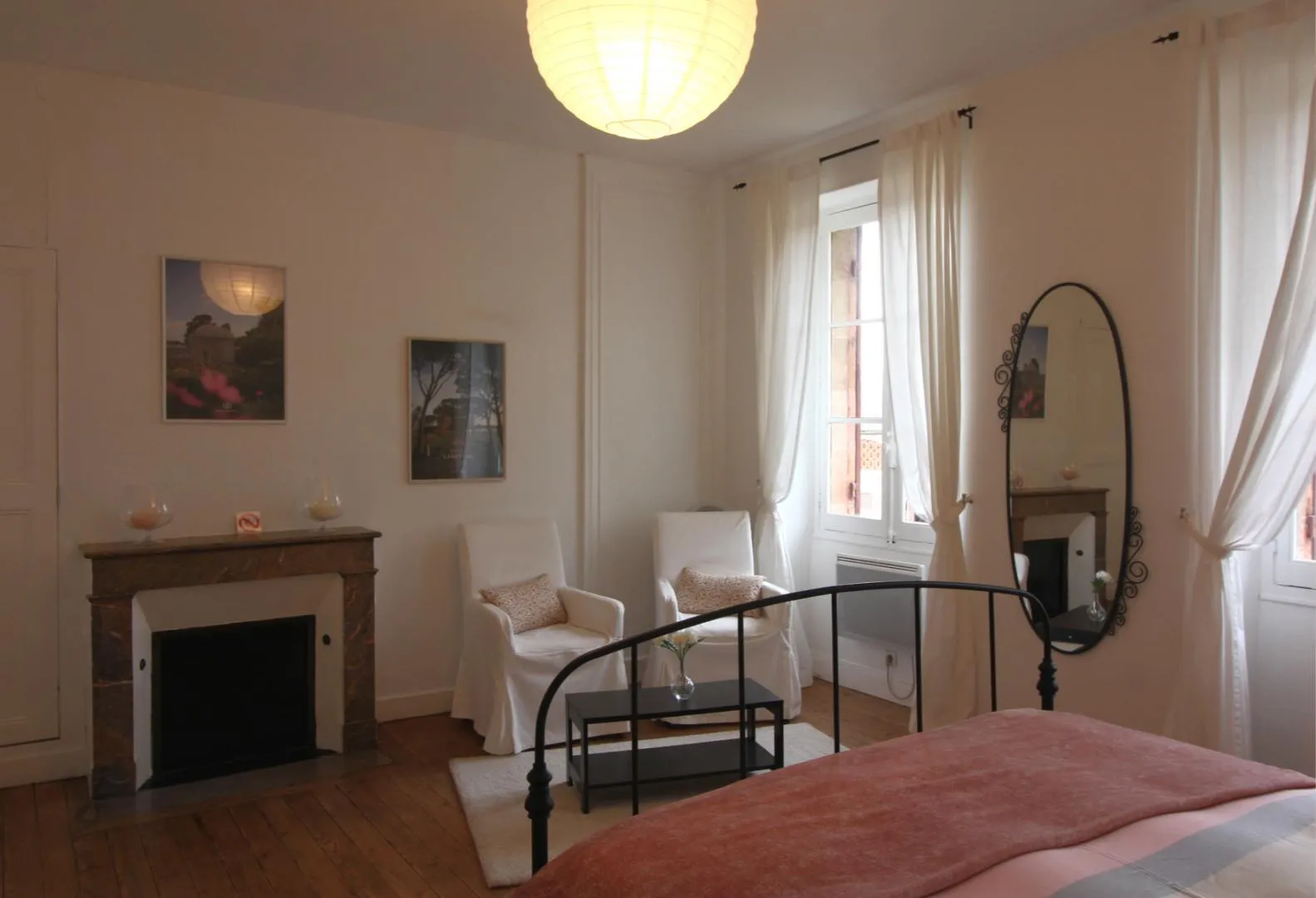 Elegant bedroom at the Maison des Hôtesses in Bordeaux featuring a wrought iron bed, wooden fireplace, white armchairs, ornate mirror, and windows overlooking the city