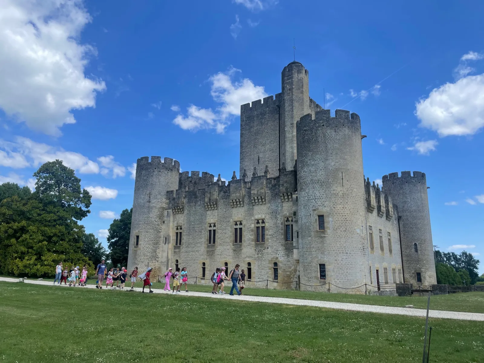 Roquetaillade Castle, a medieval fortress featuring white stone towers with crenellations, surrounded by green lawns with visitors strolling in front, located near Bordeaux.