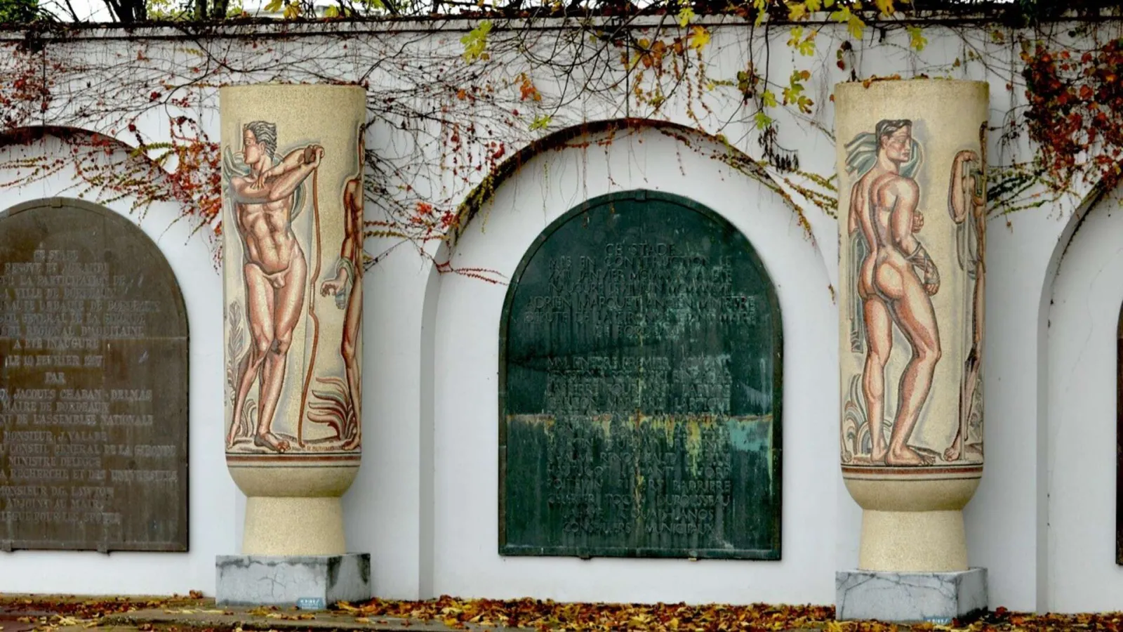 White facade with green arched entrance of Chaban-Delmas Stadium in Bordeaux, decorated with male figure reliefs and commemorative plaques, partially covered with autumn ivy.