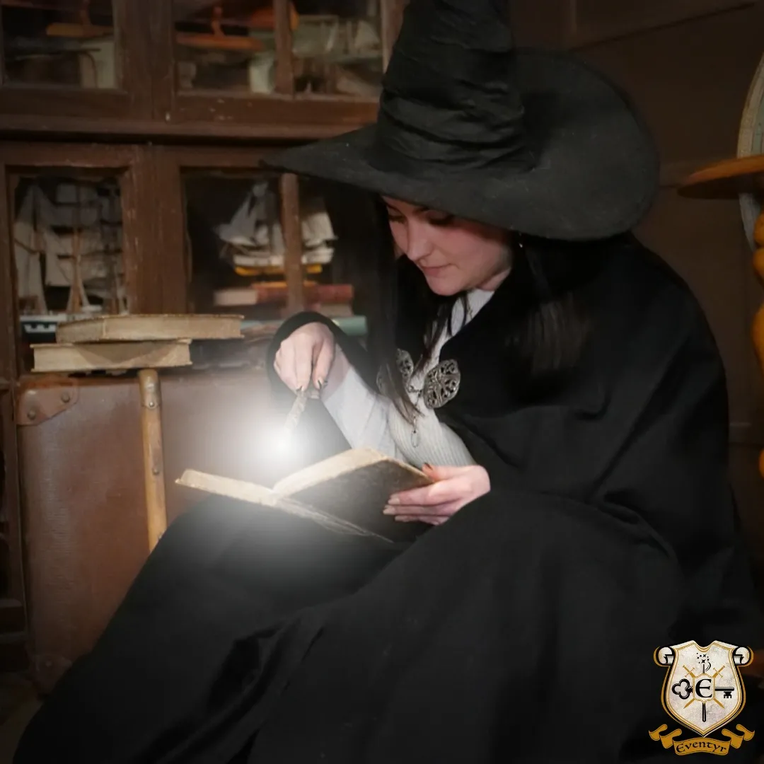 Woman dressed in black with a cowboy hat reading an open book by lamplight, seated in a rustic indoor setting in Bordeaux