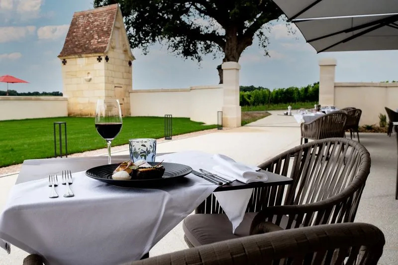 Elegant restaurant table set on a terrace overlooking vineyards, with red wine glass, black plate and rattan chair under gray parasol, historic castle in background
